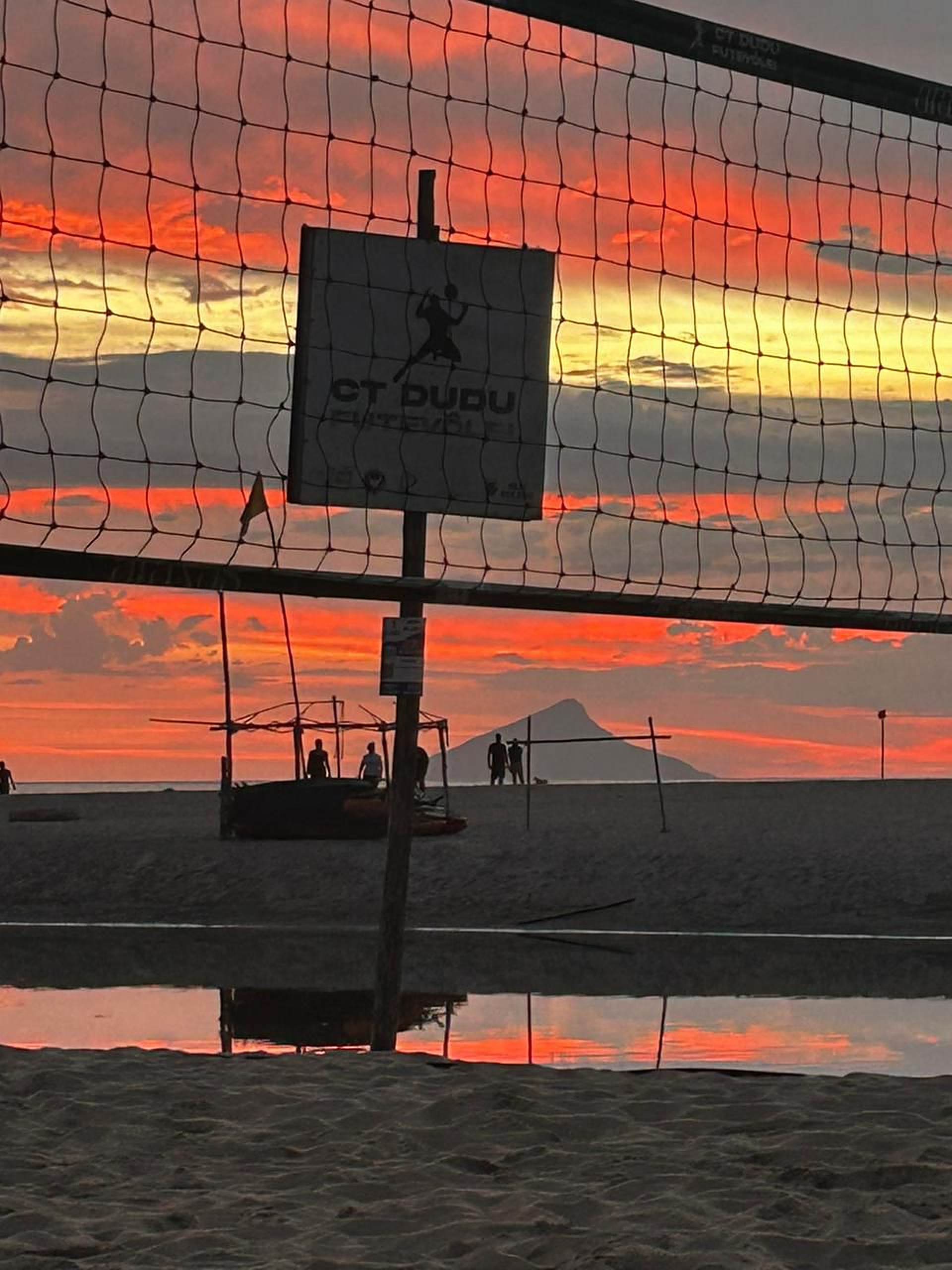 Placa do CT Dudu Futevôlei vista através da rede com céu alaranjado